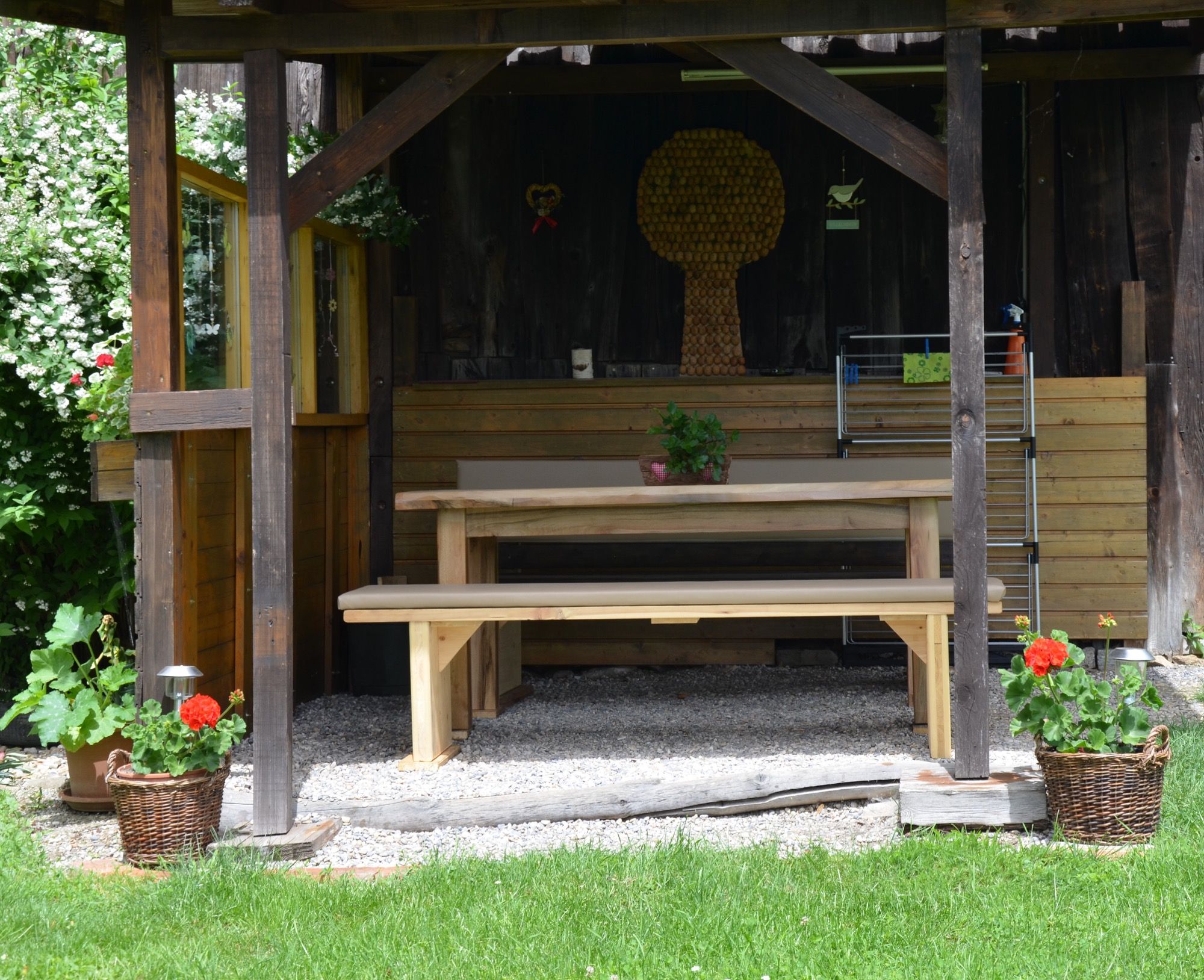 Eine rustikale Gartenlaube mit Holztisch und Bänken, umgeben von Pflanzen und Blumen in Körben.
