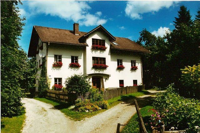Ein traditionelles weißes Haus mit roten Fensterläden und Blumenkästen, umgeben von Bäumen und einem Garten, unter blauem Himmel.