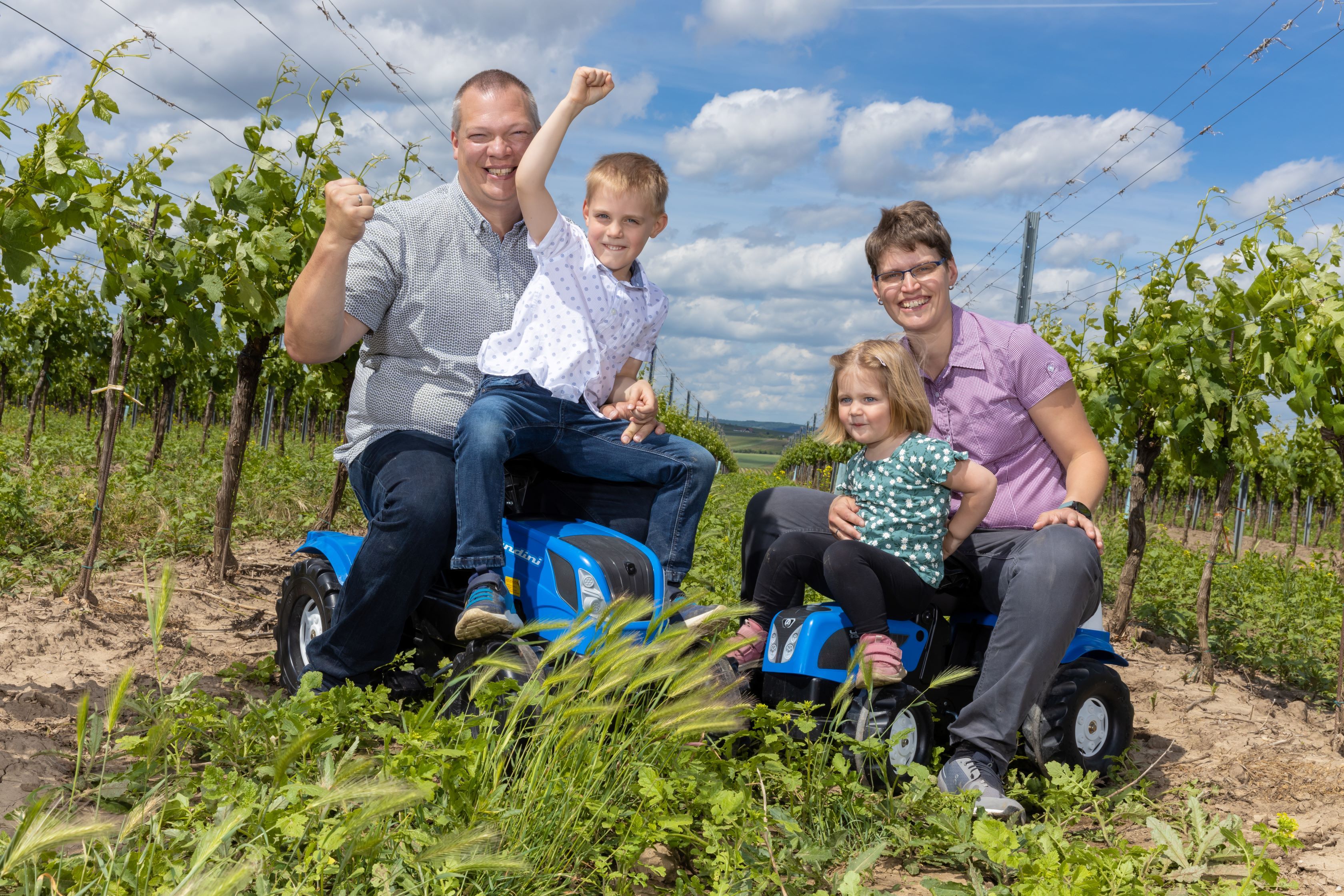 Familie sitzt auf Spielzeugtraktoren im Weinberg.