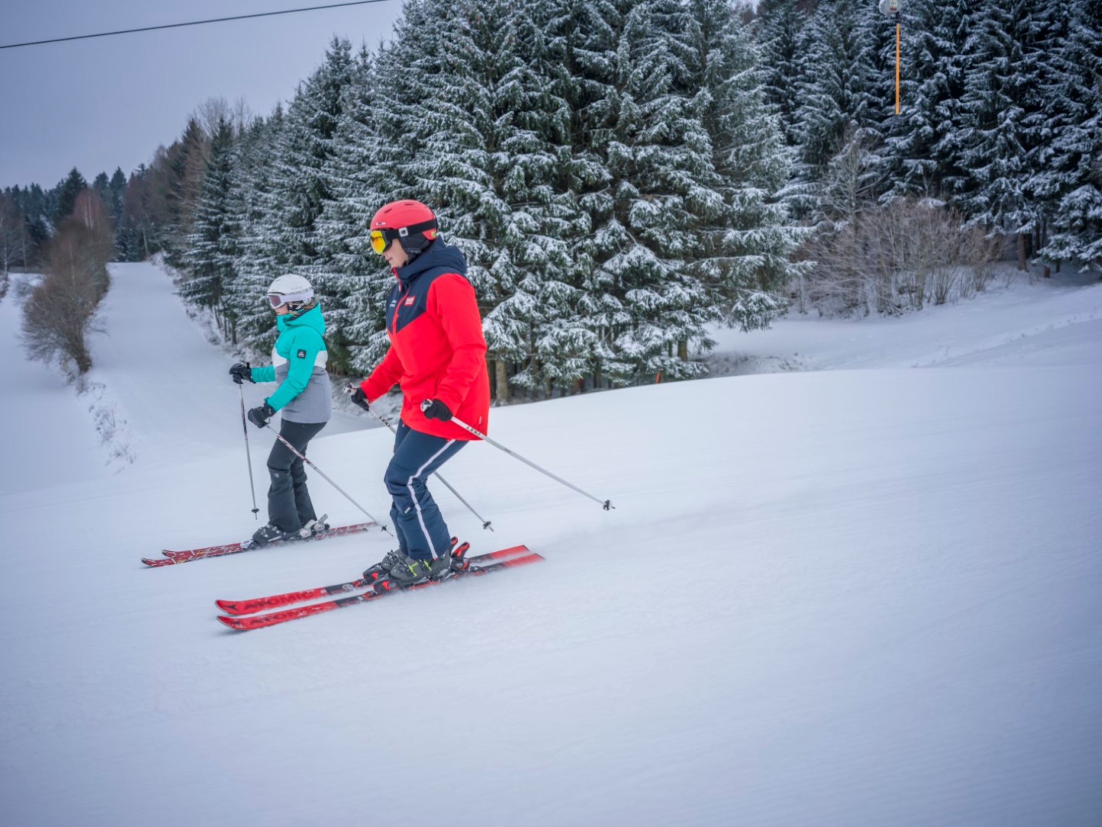 Zwei Skifahrer auf einer verschneiten Piste, umgeben von schneebedeckten Bäumen.