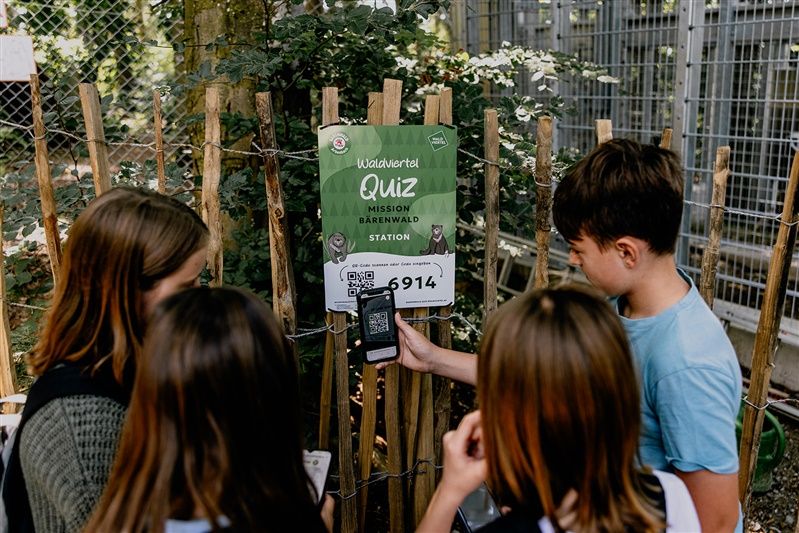Kinder stehen vor einem Schild mit der Aufschrift 'Waldviertel Quiz Mission Bärenwald Station' und scannen einen QR-Code mit einem Smartphone.