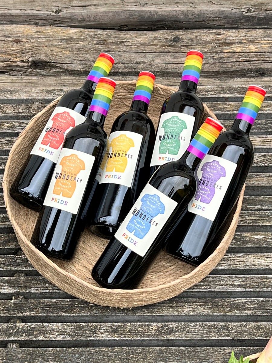 Sechs Weinflaschen mit Regenbogenetiketten in einem Korb auf Holztisch.