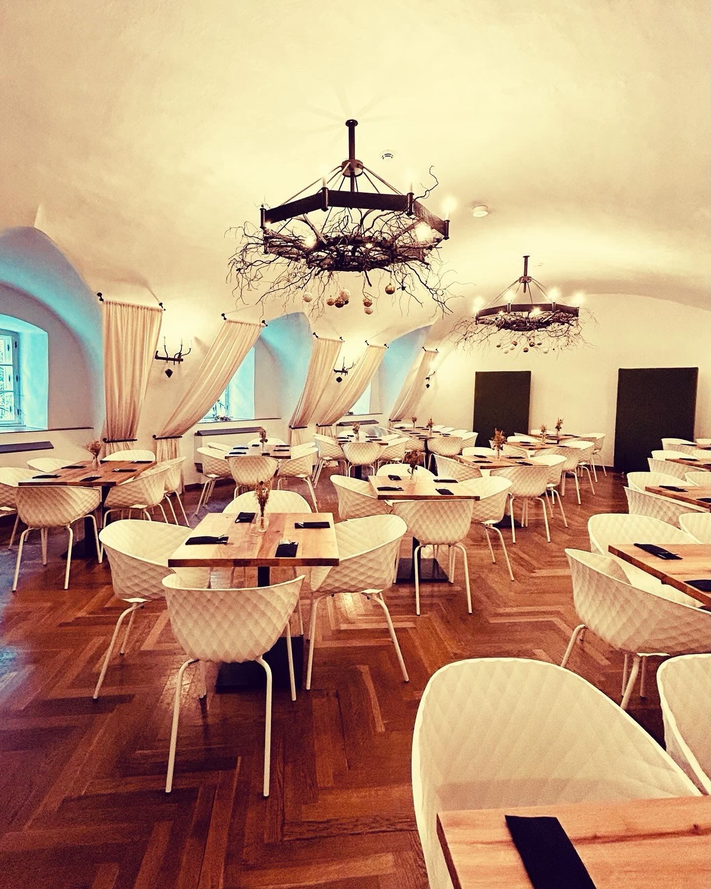 Eleganter Speisesaal mit Parkettboden, weißen Stühlen, Holztischen und dekorativen Kronleuchtern.