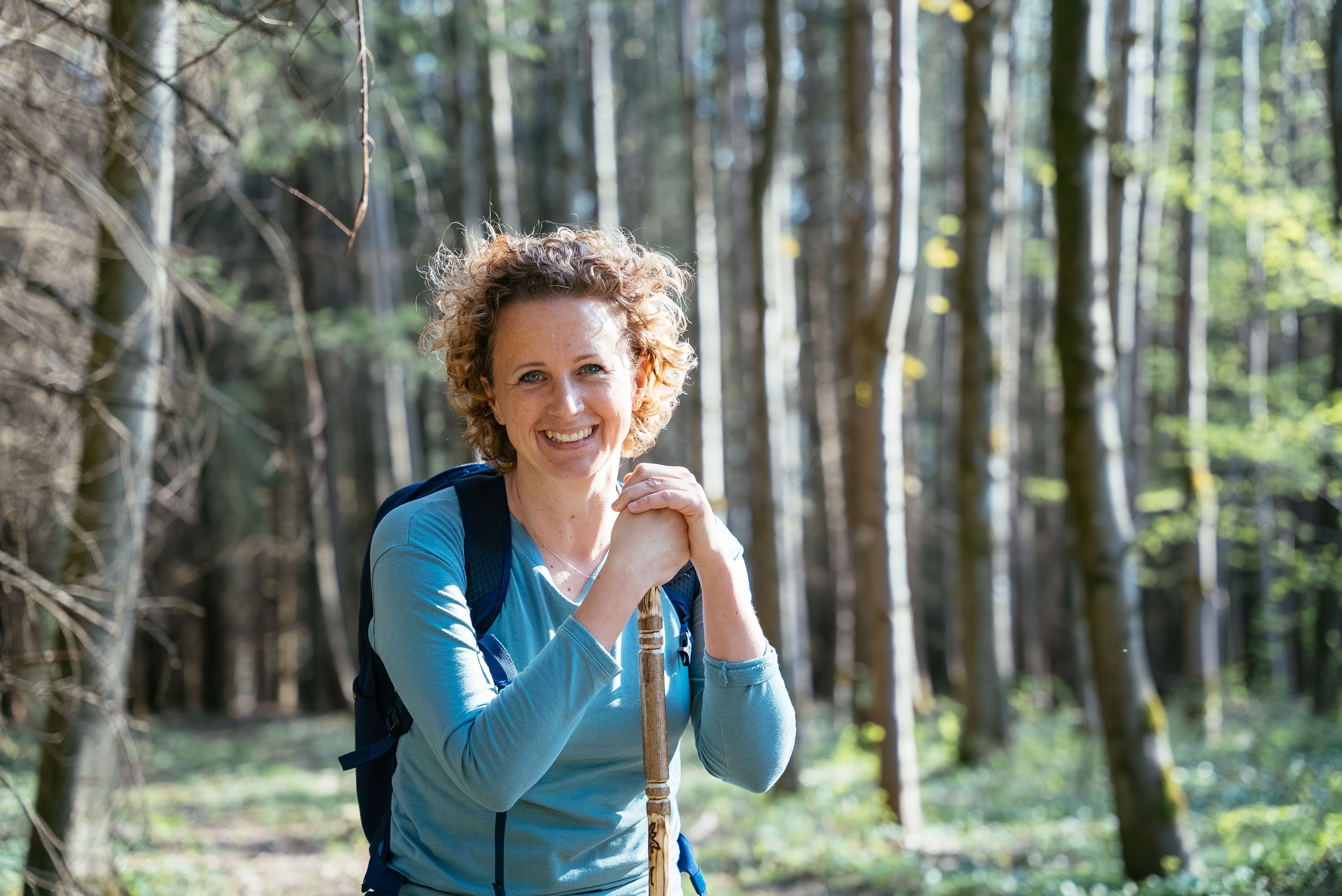 Wanderin mit Rucksack und Wanderstock lächelt in einem sonnigen Wald in die Kamera.