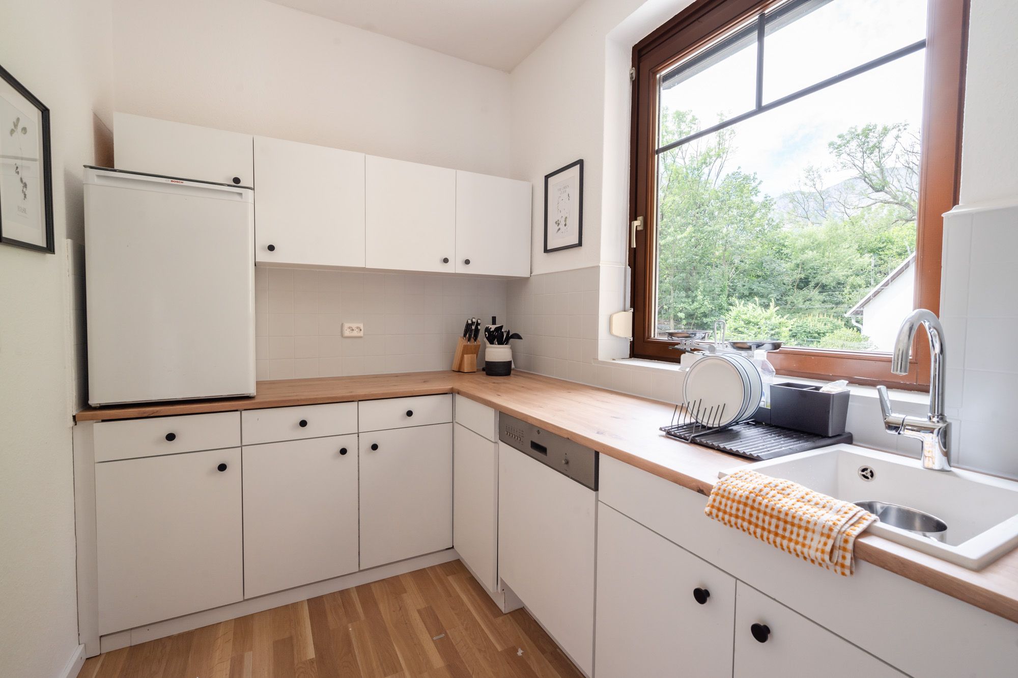 Moderne Küche mit weißen Schränken, Holzarbeitsplatte und großem Fenster mit Blick ins Grüne.