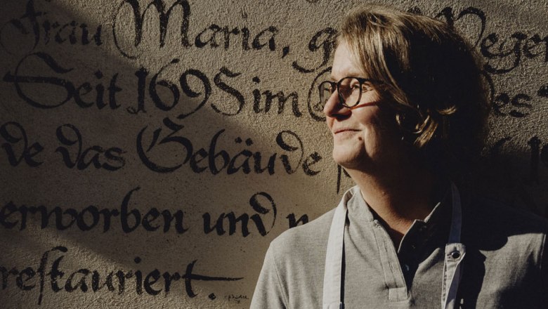 Person vor historischer Wand mit alter Schrift, l&auml;chelt im Sonnenlicht.