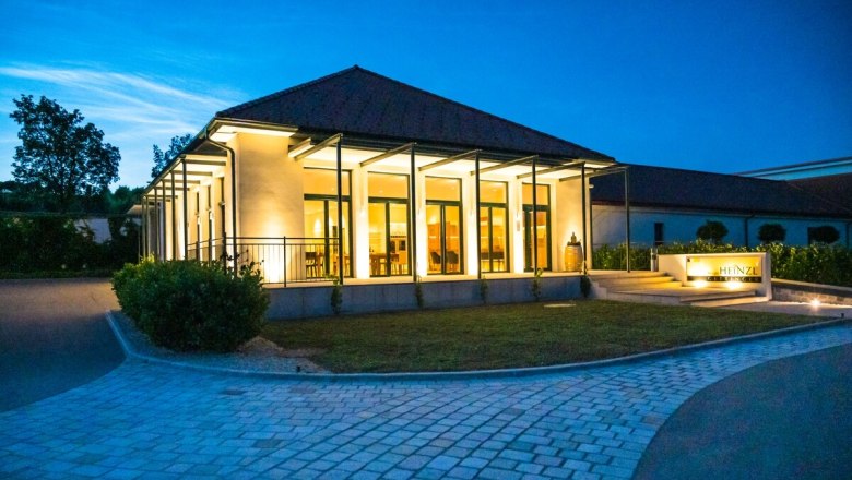 Ein modernes Geb&auml;ude mit Glasfronten, beleuchtet in der D&auml;mmerung, umgeben von einem gepflasterten Weg und Gr&uuml;nfl&auml;chen.