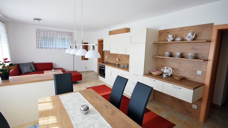 Moderner Wohn- und Essbereich mit offener K&uuml;che, rotem Sofa, Holztisch und wei&szlig;en H&auml;ngelampen.