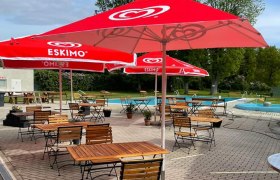 Ein Freibad mit roten Sonnenschirmen und leeren Holztischen auf einer gepflasterten Terrasse.