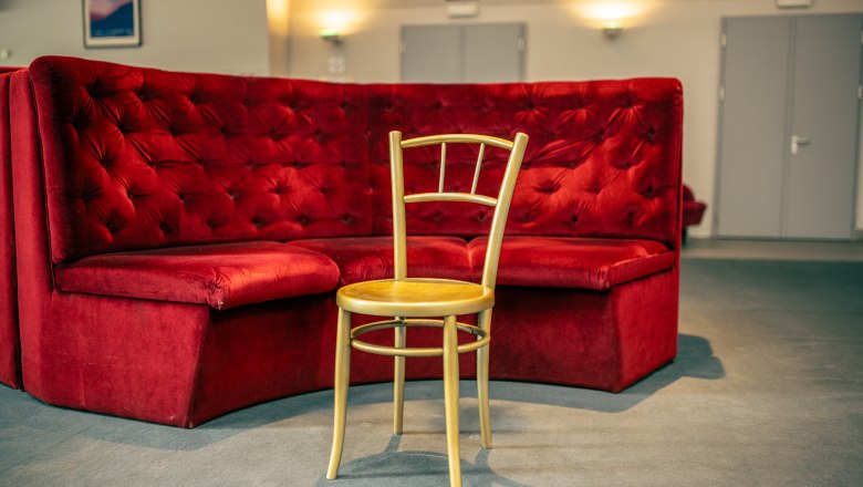 Ein einzelner Holzstuhl steht vor einer roten Samtbank in einem Theaterfoyer.