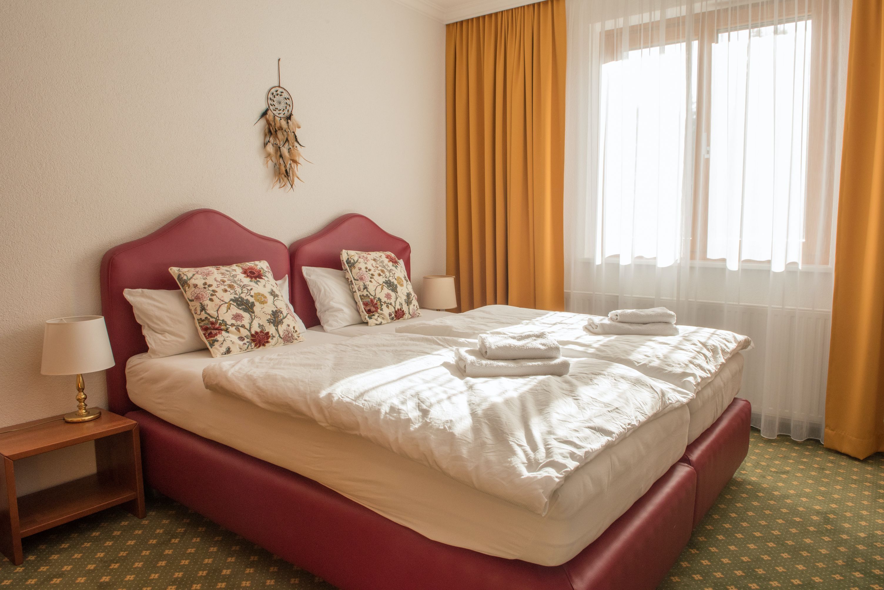 Ein gemütliches Doppelzimmer mit zwei Betten, gelben Vorhängen und einem Traumfänger an der Wand.