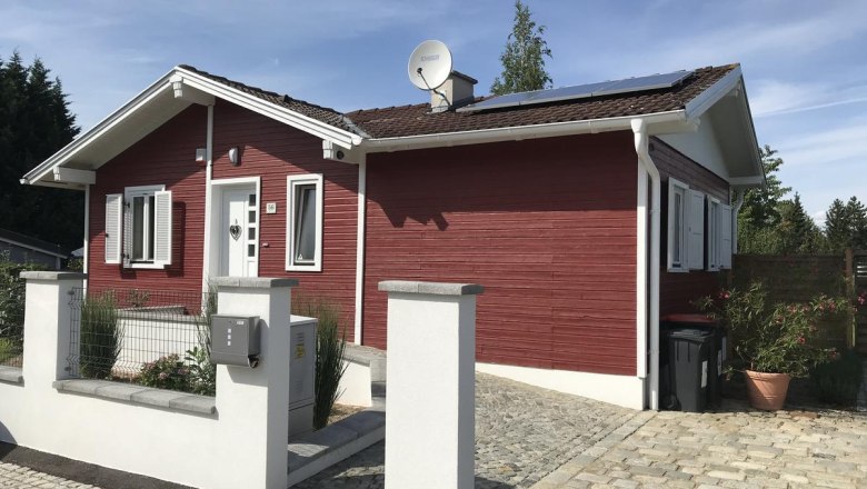 Rotes Holzhaus mit weißen Fensterläden und Solarpanelen auf dem Dach, umgeben von einem gepflasterten Hof und einer niedrigen Mauer.