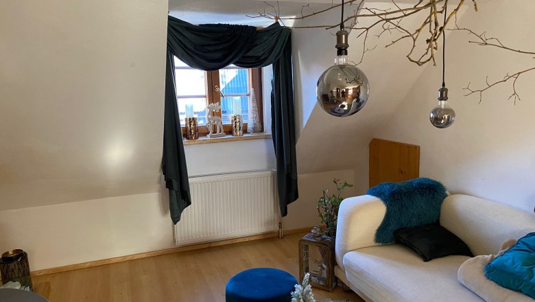 Gemütliches Wohnzimmer mit Sofa, Holzboden, Fenster mit Vorhängen, dekorativen Kerzen und hängenden Lampen.