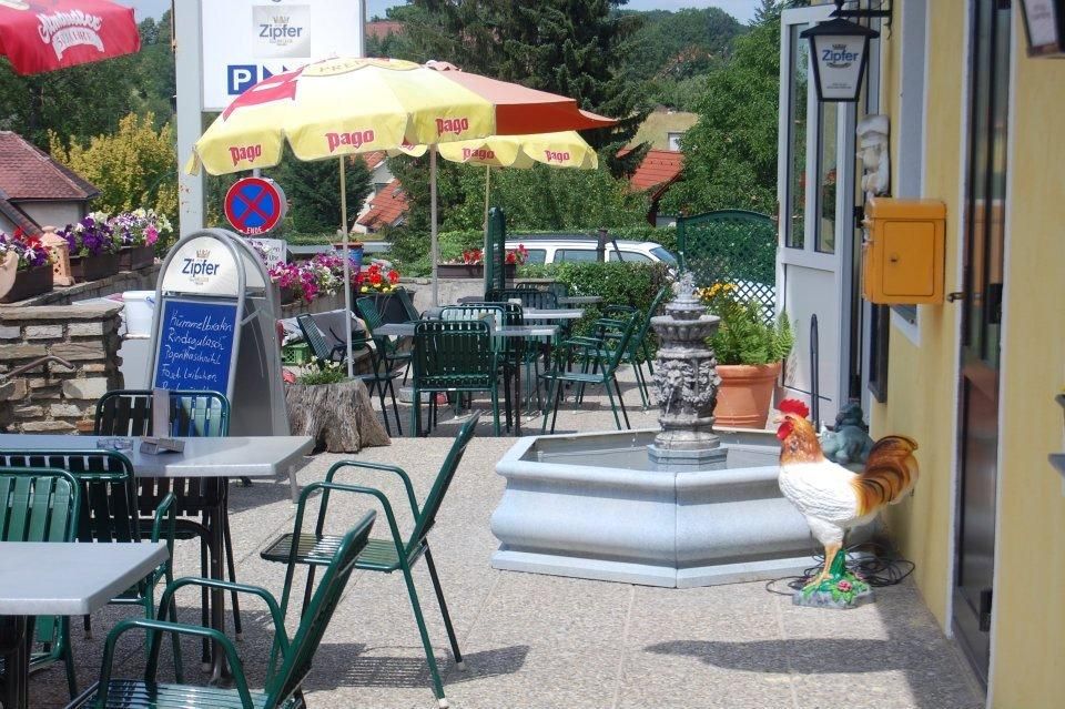 Ein Gastgarten mit Tischen, Stühlen, Sonnenschirmen und einem Brunnen. Ein Hahn steht neben dem Brunnen.