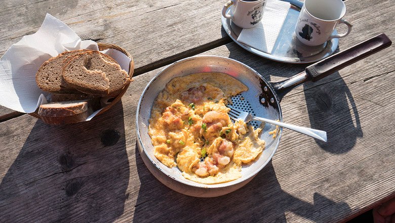 Pfanne mit R&uuml;hrei und Speck auf Holztisch, daneben Brotkorb und zwei Tassen.