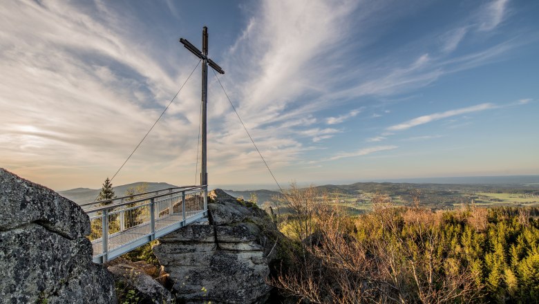 Ein Gipfelkreuz auf einem Felsen mit einer Aussichtsplattform, umgeben von Wald und einem weiten Blick &uuml;ber die Landschaft unter einem blauen Himmel.