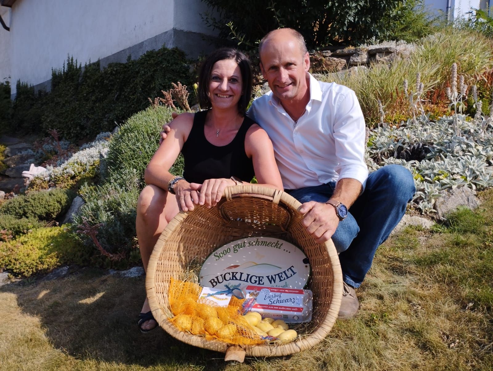 Eine Frau und ein Mann sitzen lächelnd in einem Garten und halten einen Korb mit Kartoffeln und Eiern.