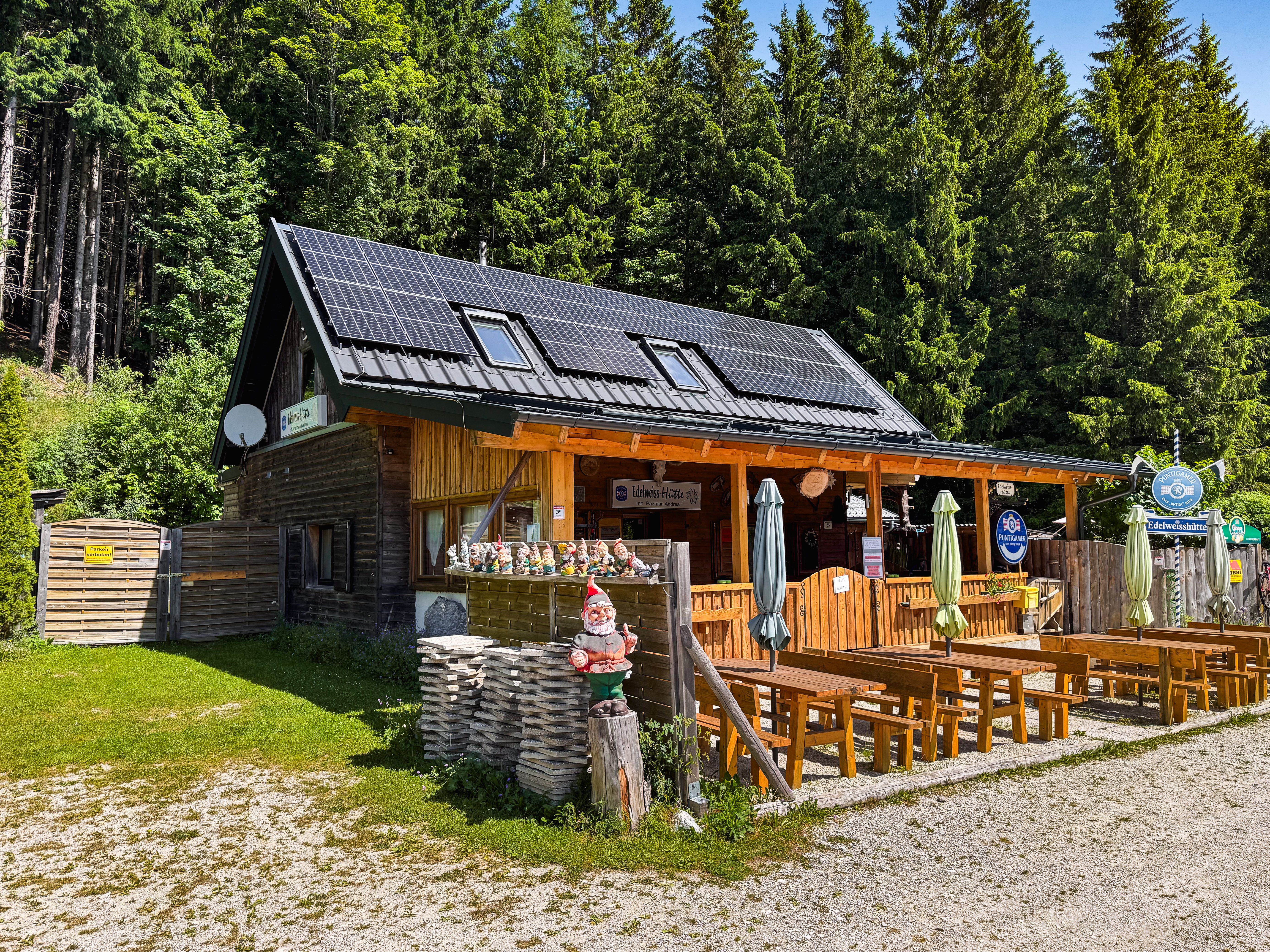 Eine Berghütte mit Solarpanelen auf dem Dach, umgeben von Bäumen. Vor der Hütte stehen Holzbänke und ein Gartenzwerg.
