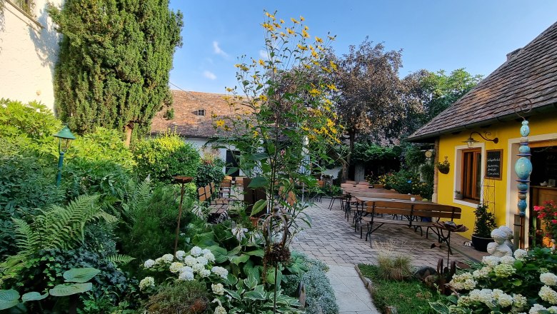 Ein gem&uuml;tlicher Garten mit gelbem Geb&auml;ude, Holzb&auml;nken und &uuml;ppiger Vegetation unter blauem Himmel.