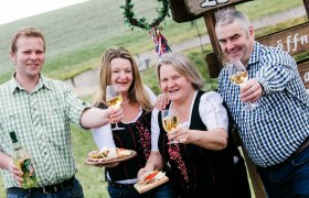 Vier Personen sto&szlig;en mit Weingl&auml;sern an, im Freien vor einem Schild.