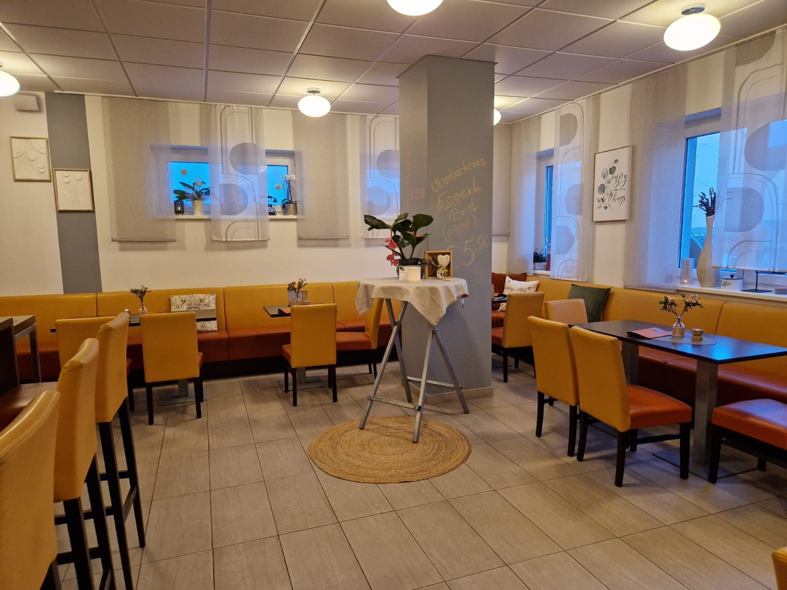 Innenansicht eines gemütlichen Cafés mit gelben Stühlen und Tischen, dekoriert mit Pflanzen und Bildern an den Wänden.