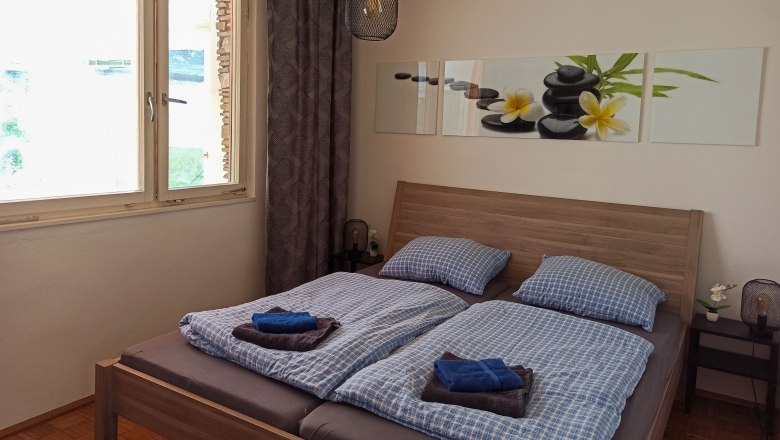 Schlafzimmer mit Doppelbett, blauer Bettw&auml;sche, Bild mit Steinen und Blumen an der Wand, Fenster und Nachttischlampe.