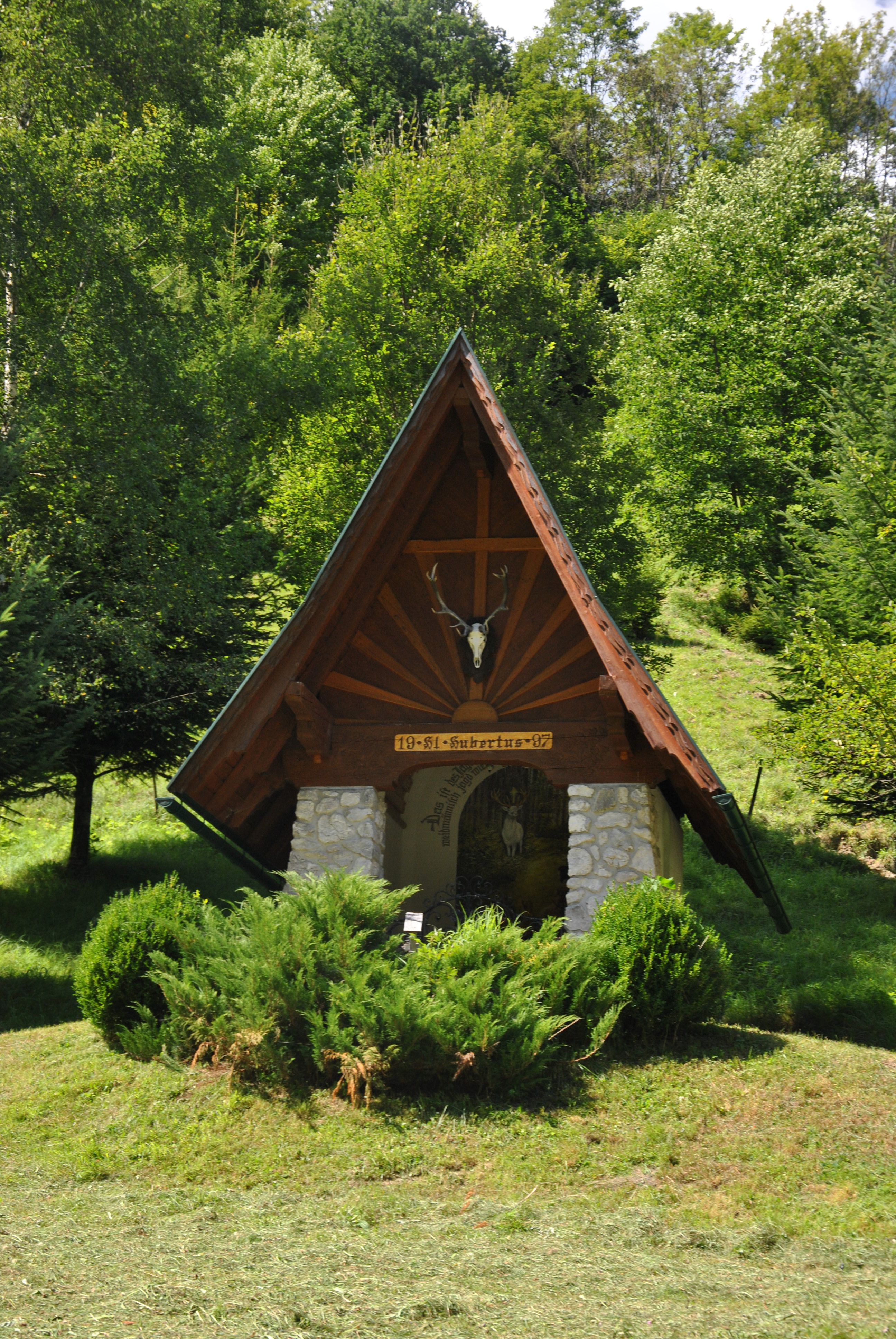 Eine kleine Kapelle mit spitzem Dach und Hirschgeweih, umgeben von Bäumen und Büschen.