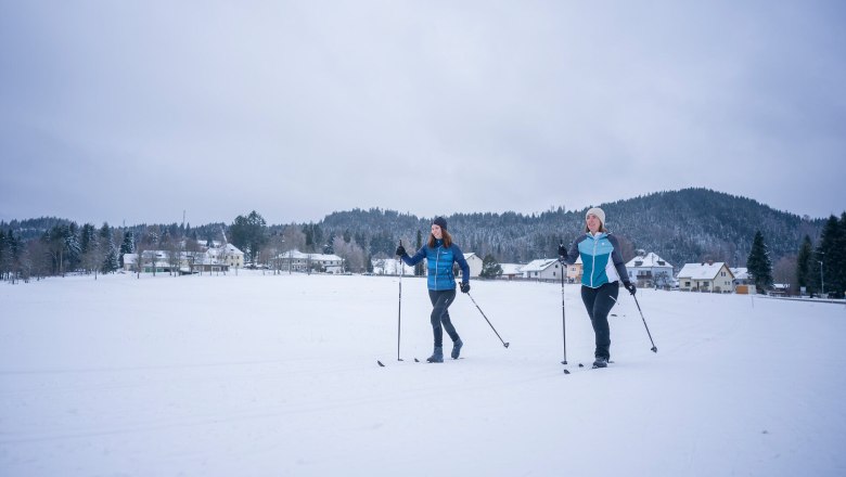 Zwei Personen beim Langlaufen auf einer schneebedeckten Fl&auml;che vor einem Dorf und bewaldeten H&uuml;geln.