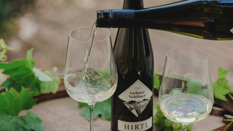 Wei&szlig;wein wird in ein Glas gegossen, daneben eine Flasche Gr&uuml;ner Veltliner von Hirtl und gr&uuml;ne Trauben auf einem Holztisch.