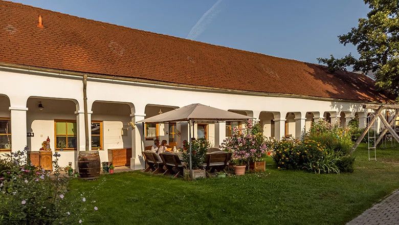 Ein traditionelles Weingut mit weißen Arkaden, einem roten Ziegeldach und einem Garten mit Sitzbereich und Blumen.