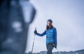 Eine Frau beim Langlaufen auf einer schneebedeckten Fl&auml;che mit H&auml;usern im Hintergrund.