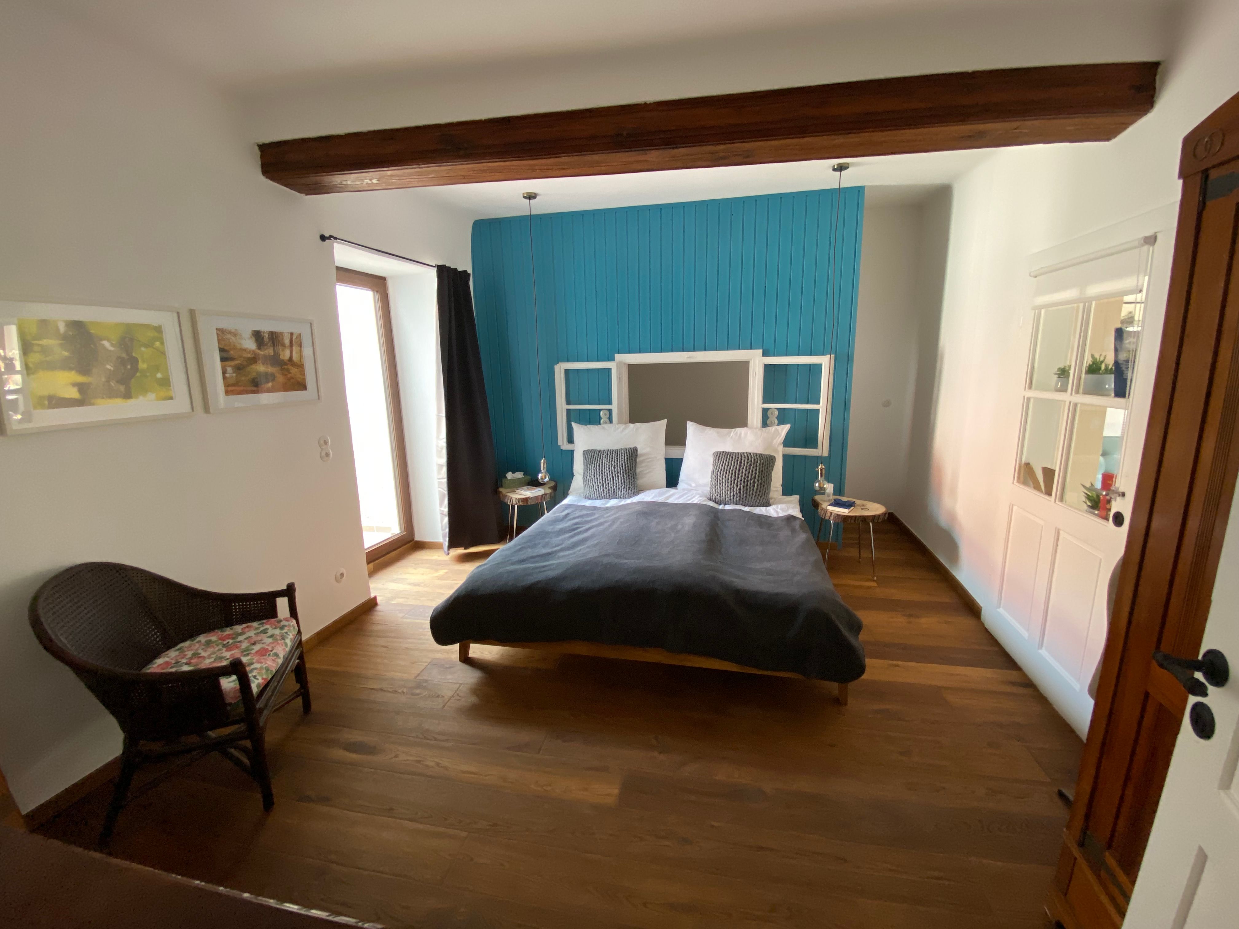 Modernes Schlafzimmer mit Holzboden, blauem Akzent an der Wand, Doppelbett und Rattan-Stuhl.