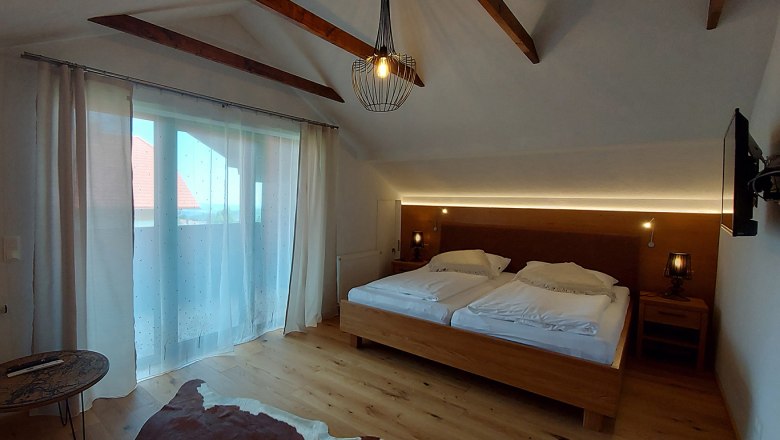 Ein gem&uuml;tliches Doppelzimmer mit Holzboden, einem gro&szlig;en Bett, Vorh&auml;ngen und einem kleinen Tisch. Eine Lampe h&auml;ngt von der Decke.