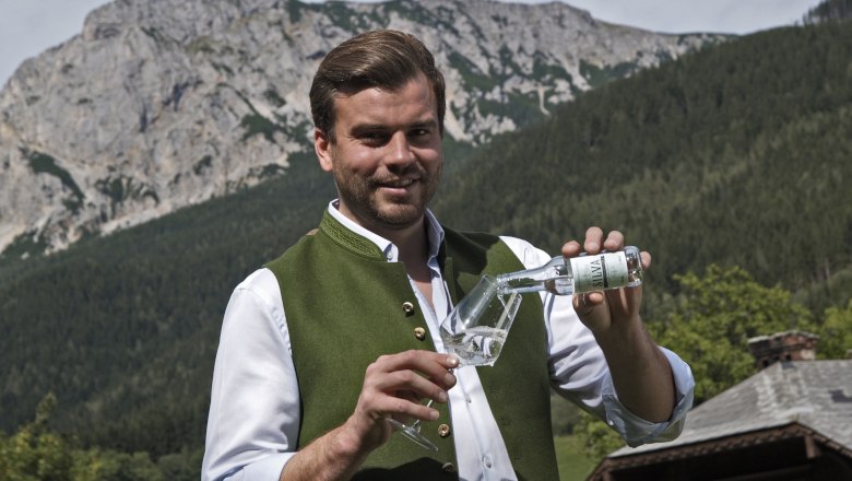 Mann in traditioneller Kleidung schenkt ein Getr&auml;nk in ein Glas ein, mit Berglandschaft im Hintergrund.