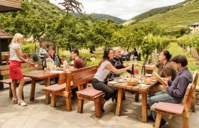 Menschen sitzen an Holztischen im Freien, umgeben von Weinbergen und B&auml;umen, w&auml;hrend eine Kellnerin Getr&auml;nke serviert.