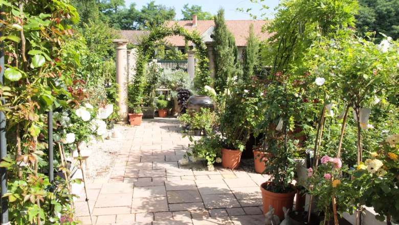 Ein gepflegter Gartenweg mit Topfpflanzen und Blumen, flankiert von Säulen und einem Torbogen, führt zu einem Gebäude mit Ziegeldach.