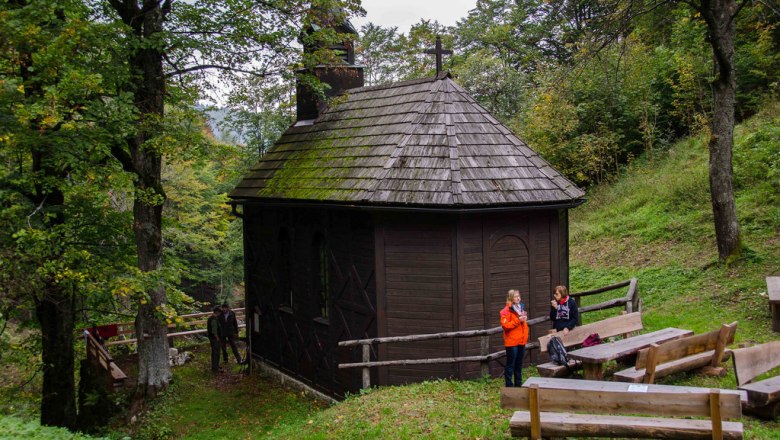 Eine kleine, hölzerne Kirche im Wald mit zwei Personen und Holzbänken im Vordergrund.
