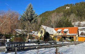 Ein Haus mit rotem Dach in einer verschneiten Landschaft, umgeben von B&auml;umen und einem Holzzaun.