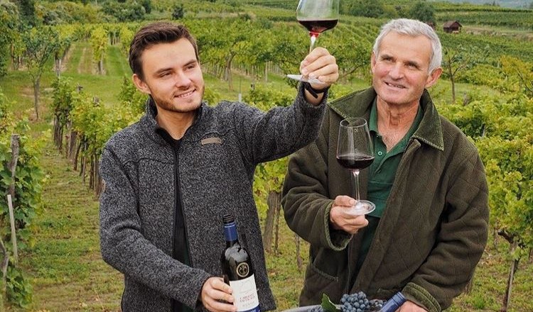 Zwei M&auml;nner in einem Weinberg halten Weingl&auml;ser mit Rotwein und l&auml;cheln. Einer h&auml;lt eine Weinflasche, im Hintergrund sind Reben und H&uuml;gel zu sehen.