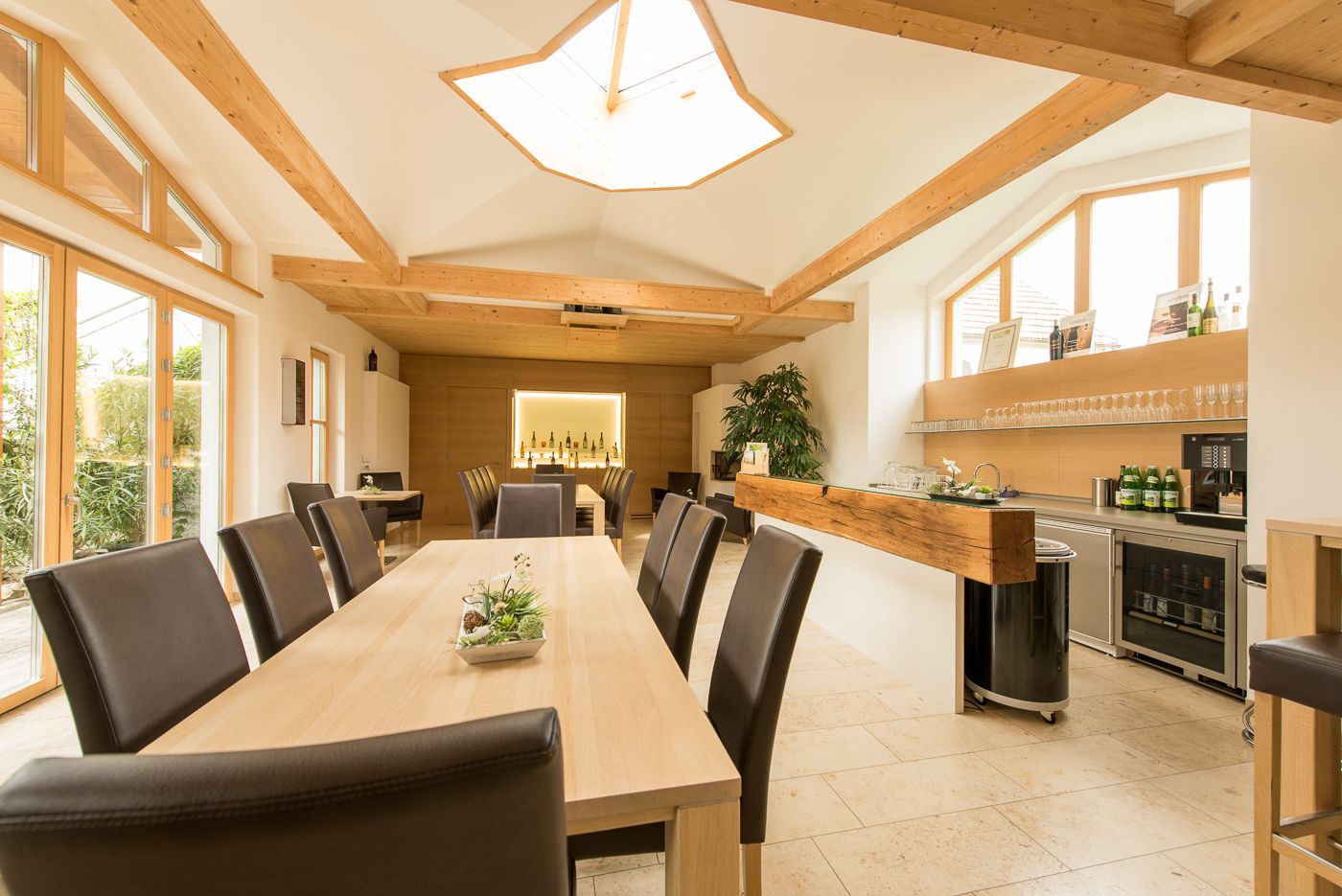 Innenansicht eines modernen Verkostungsraums mit Holztischen und -stühlen, großen Fenstern und einer Bar mit Weinflaschen.