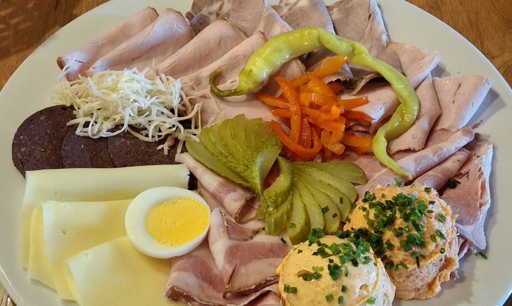 Ein Jausenteller mit verschiedenen Wurst- und K&auml;sesorten, Ei, Paprika, Gurken und Aufstrich auf einem Holztisch.