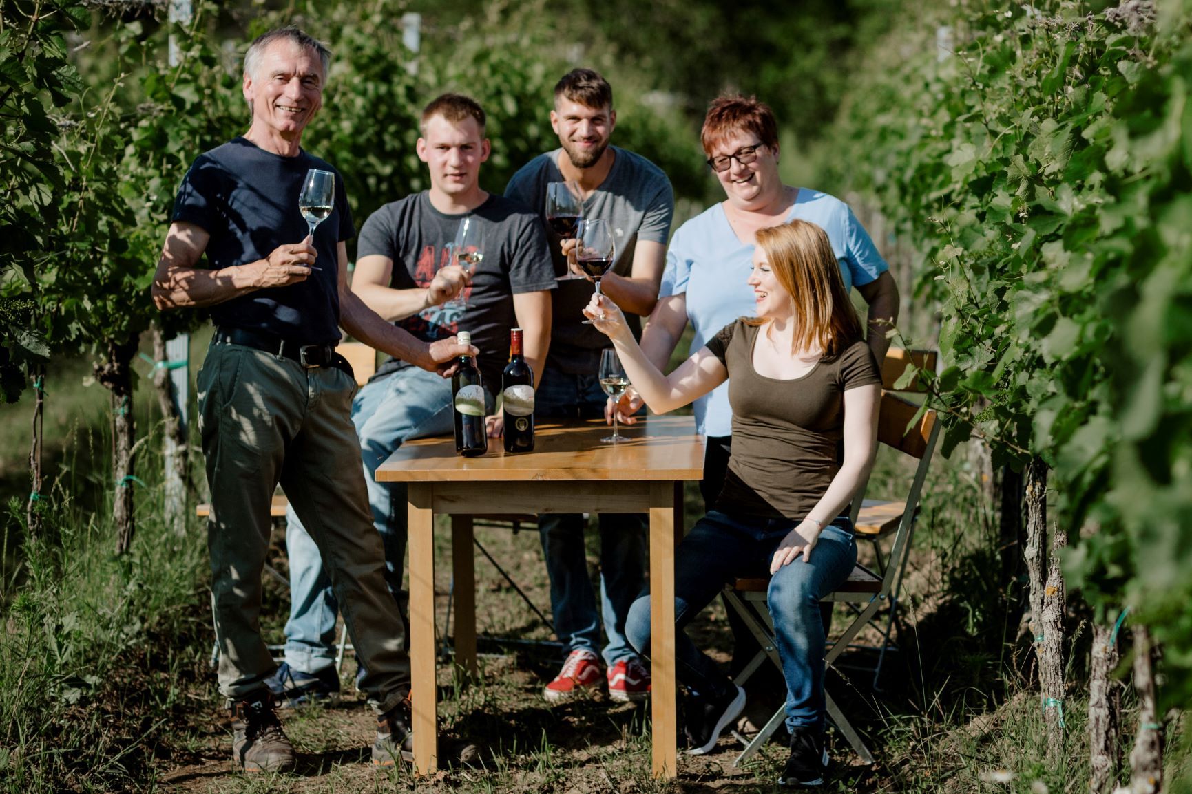Eine Gruppe von fünf Personen sitzt und steht an einem Tisch im Weinberg, hält Weingläser und lächelt.