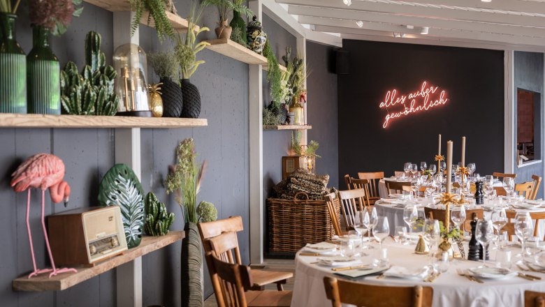 Ein stilvoll dekorierter Raum mit gedeckten Tischen, Pflanzen und einem Neon-Schriftzug an der Wand.