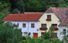 Ein traditionelles Haus mit roten Dachziegeln und blühenden Blumenkästen vor den Fenstern, umgeben von grüner Vegetation.