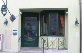 Eingangsbereich eines Geb&auml;udes mit gr&uuml;ner T&uuml;r und Fenstern, dekoriert mit Pflanzen und Laternen.