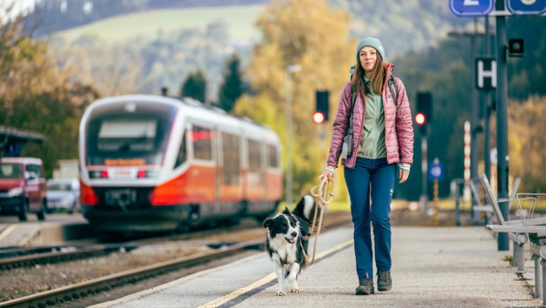 Frau mit Hund am Bahnhof Aspang, Zug im Hintergrund.