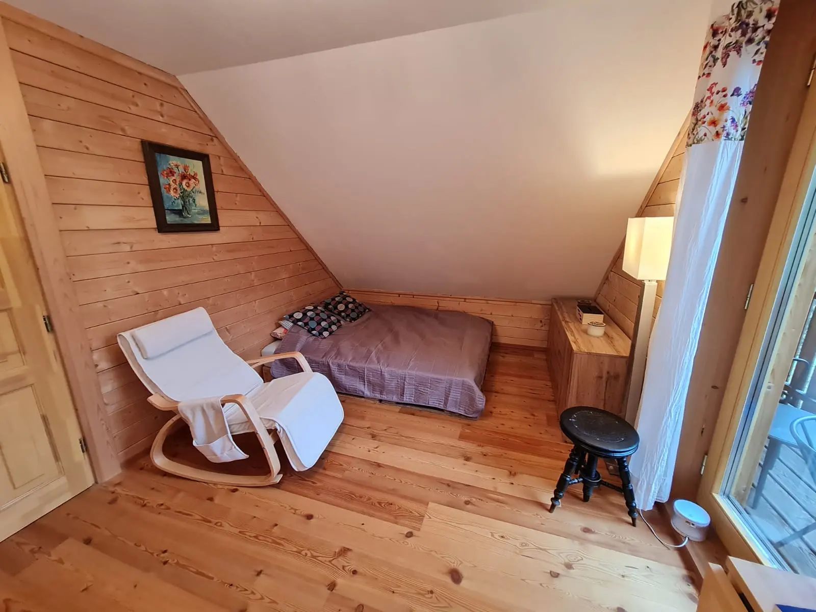 Gemütliches Zimmer mit Holzwänden, einem Bett, einem weißen Schaukelstuhl und einem kleinen Tisch.