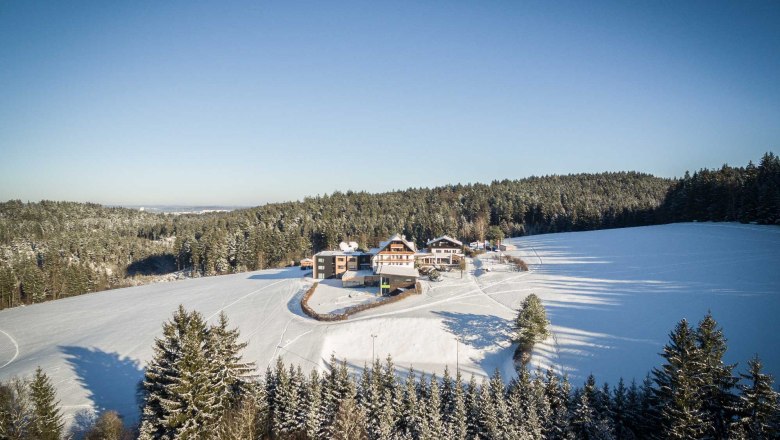 Luftaufnahme eines Hotels im Winter, umgeben von schneebedeckten Feldern und W&auml;ldern.