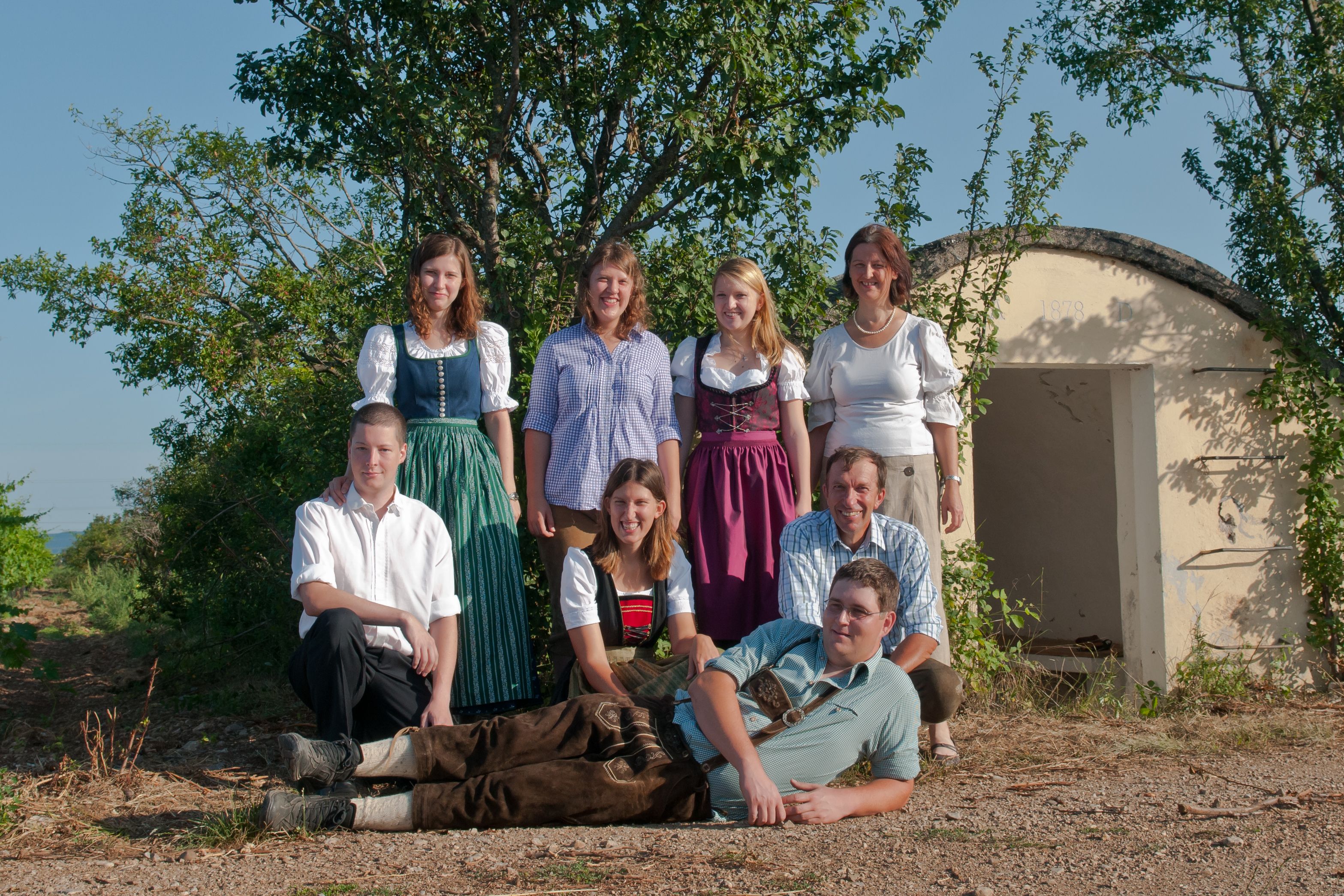 Eine Gruppe von acht Personen posiert im Freien vor einem kleinen Gebäude. Einige tragen traditionelle Kleidung.