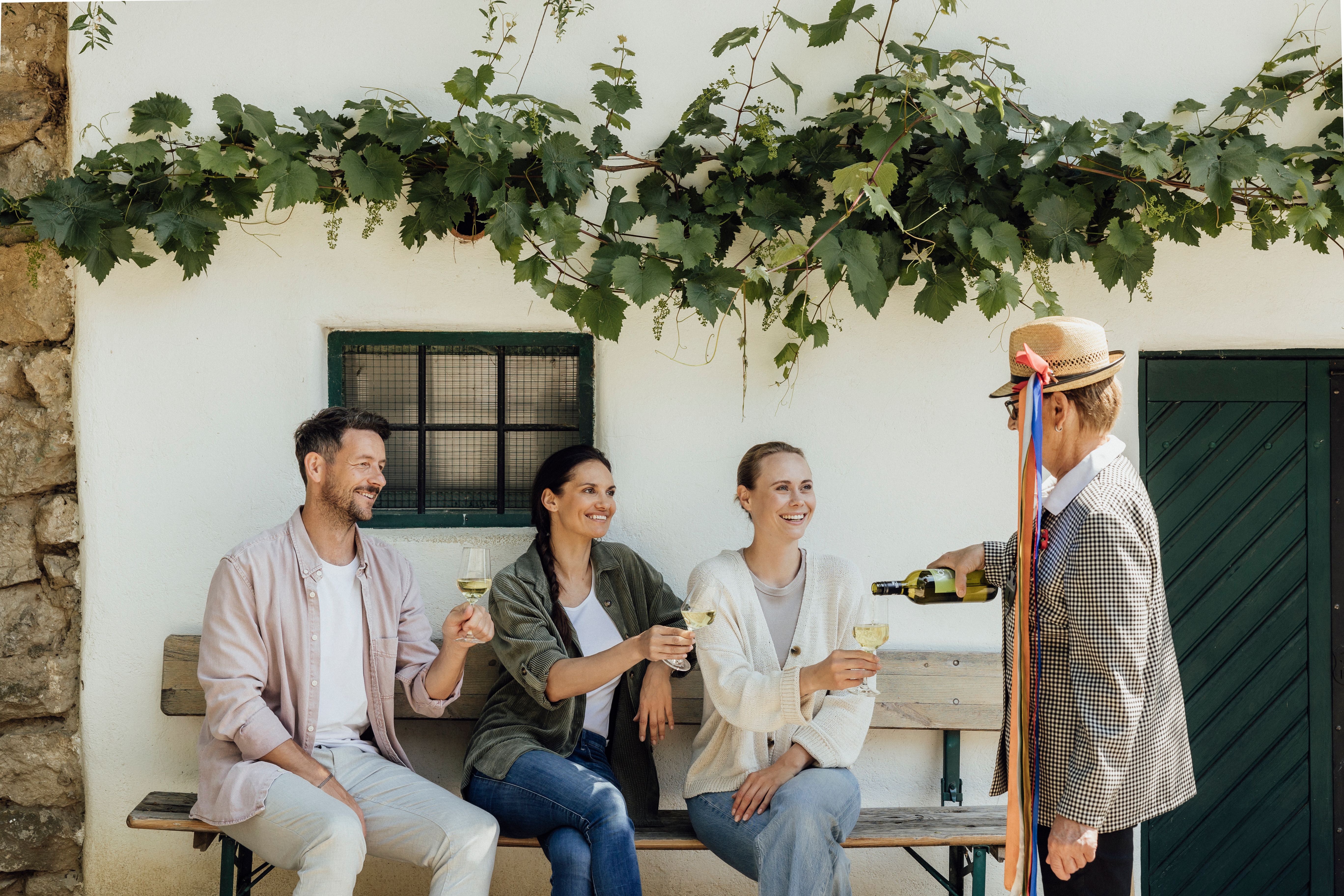 Eine Gruppe von Menschen sitzt auf einer Bank vor einem Weinkeller und genießt Wein. Eine Person schenkt Wein ein, während Weinreben die Wand schmücken.